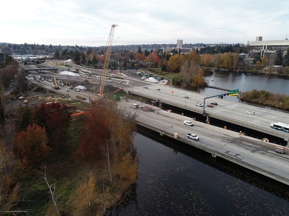 Aerial view of two bridges over water, with construction equipment at a busy interchange. Fall foliage in the foreground.