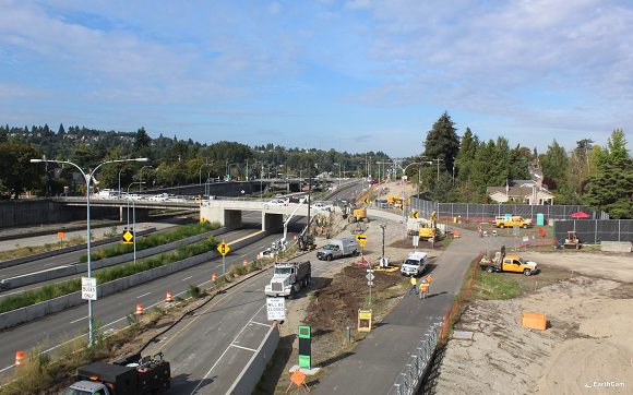 A screenshot of an online construction camera showing crews building a concrete block retaining wall on the side of a highway off-ramp.